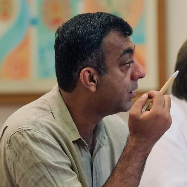 Ali Shah, President of the chapter, in a green linen shirt, raising a pencil as he speaks at a meeting table.