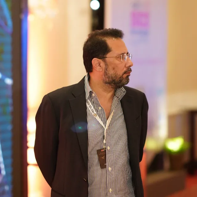 Eesa Bilal Khan, Public Relation and Member Services, in glasses, a dark blazer and a striped shirt, looking to the side at a gala.