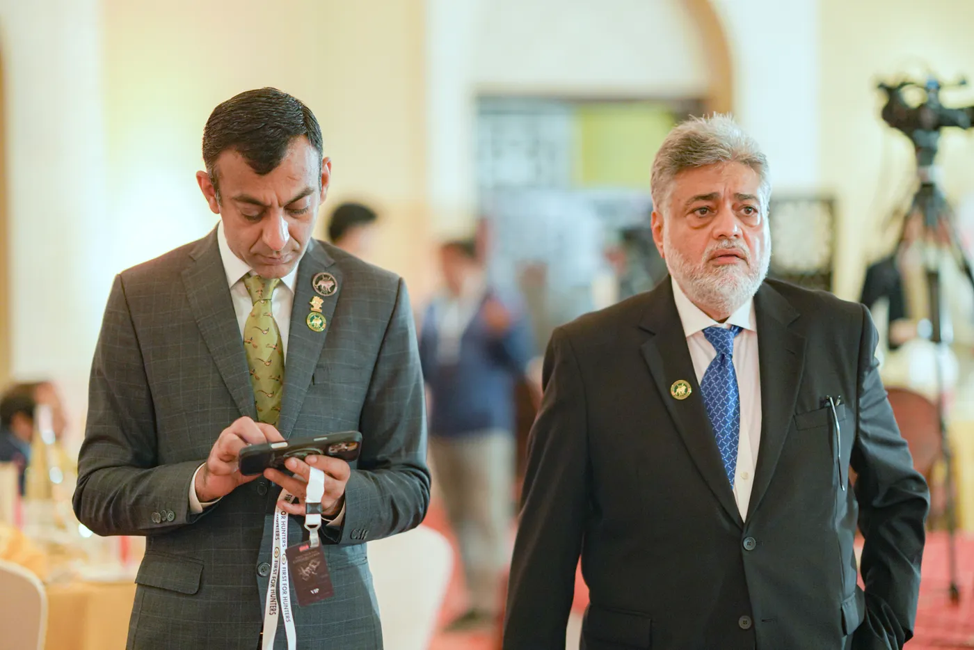 Two delegates in suits and SCI lapel pins walk through the conference hall, one checking his phone.