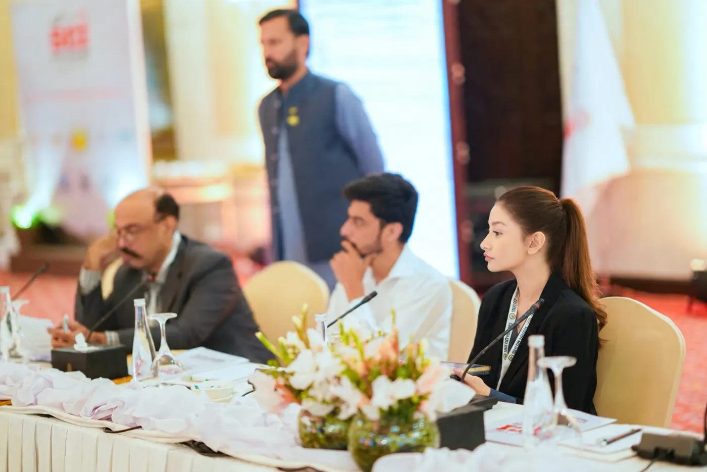 Three delegates at the conference table, with a colleague standing behind them.