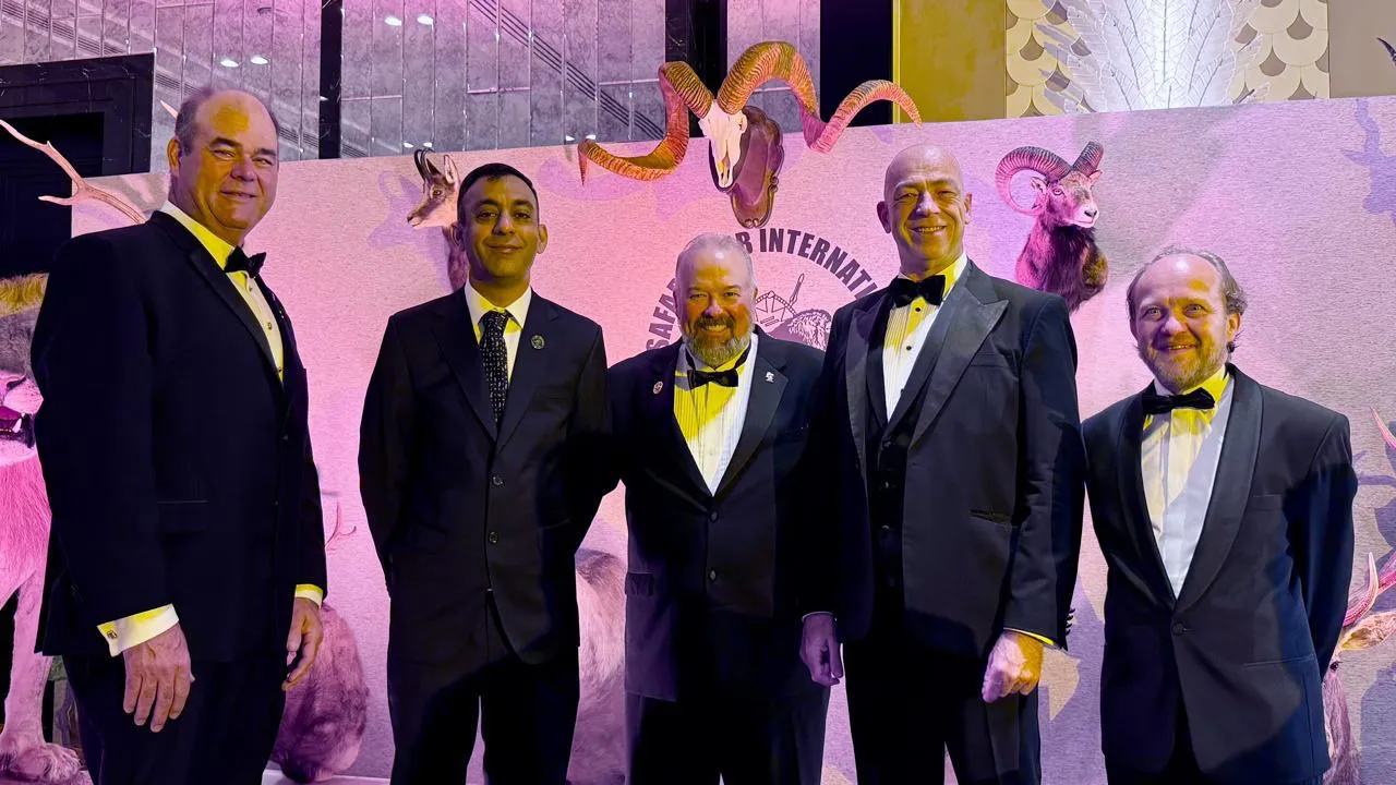 Five men in black tie stand in front of a Safari Club International backdrop of mounted sheep and deer.