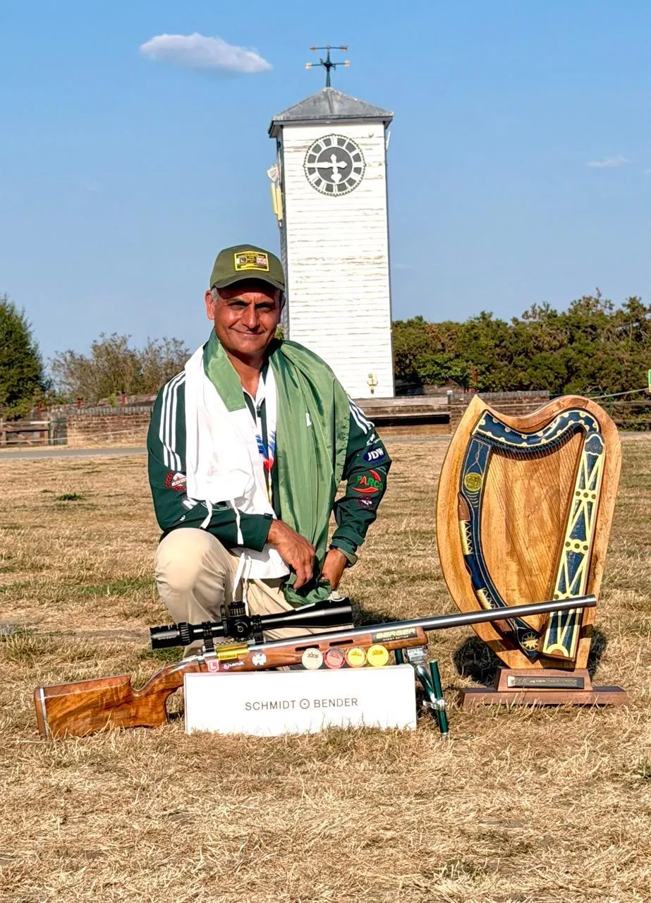 Junaid Ali Zulfiqar kneels in Pakistan colours beside his rifle and a harp-shaped trophy on a sunlit range.