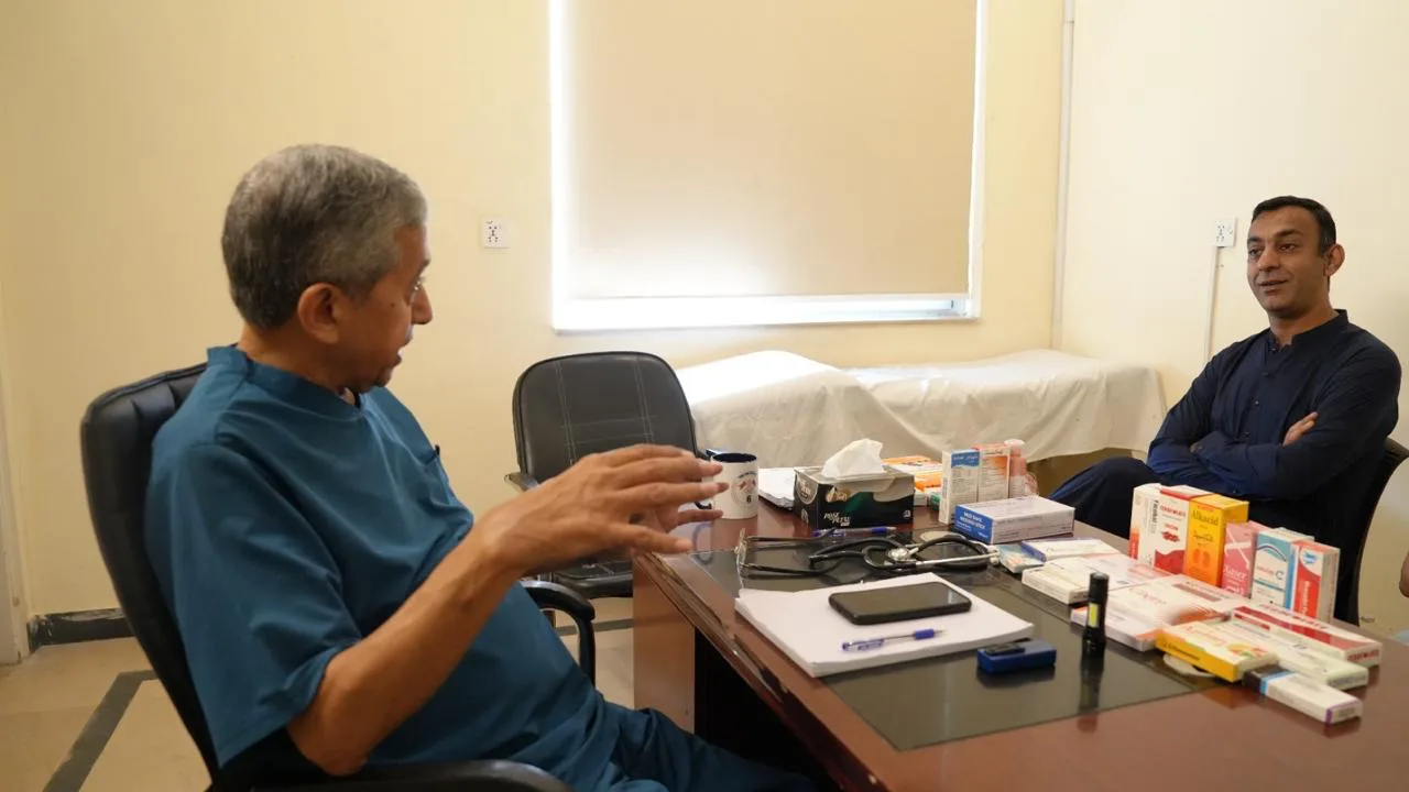 A doctor in teal scrubs talks with a visitor across a desk of prescription medicines in a consulting room.
