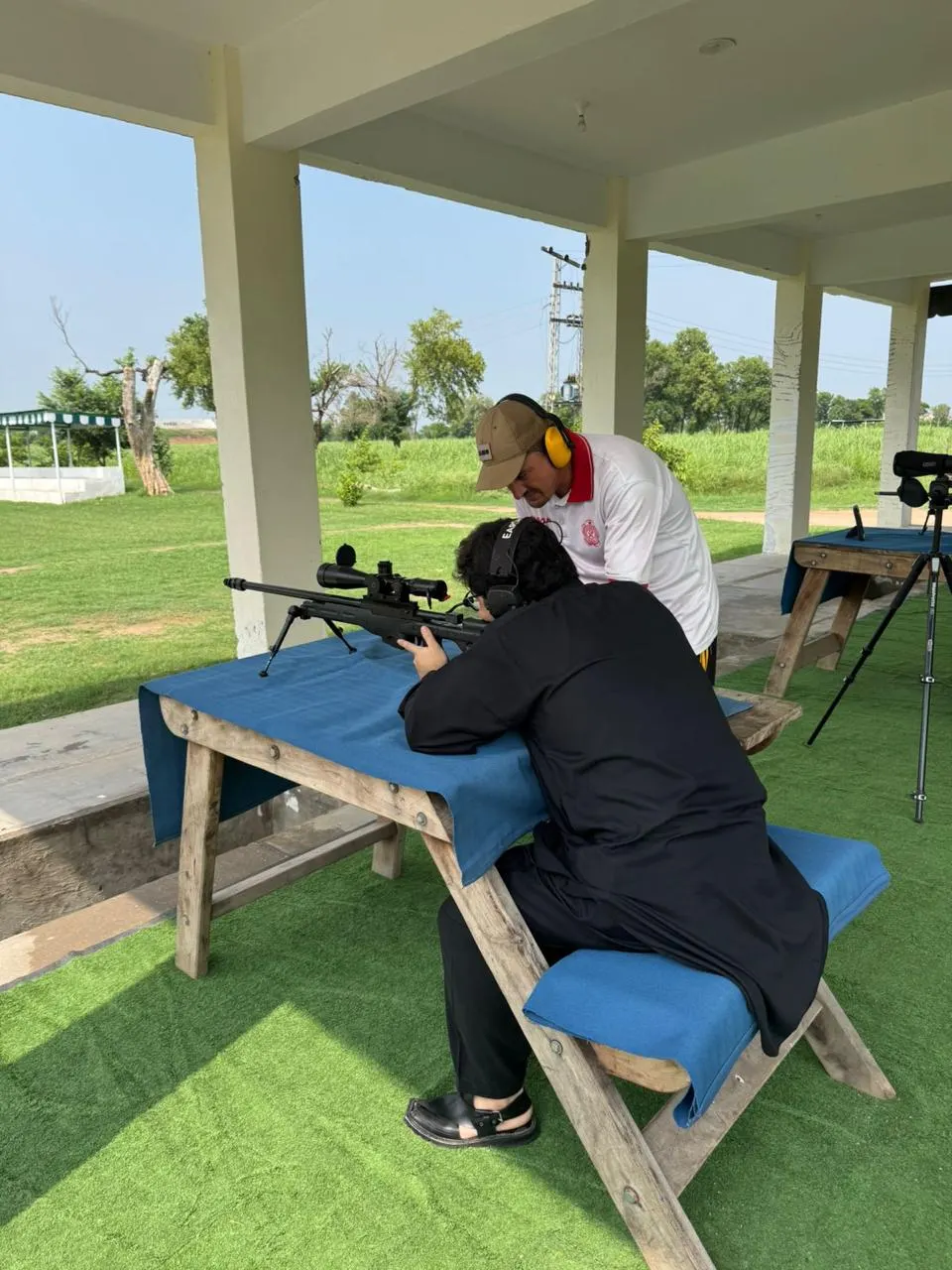 A shooter lines up a rifle at a covered range bench while an instructor watches.